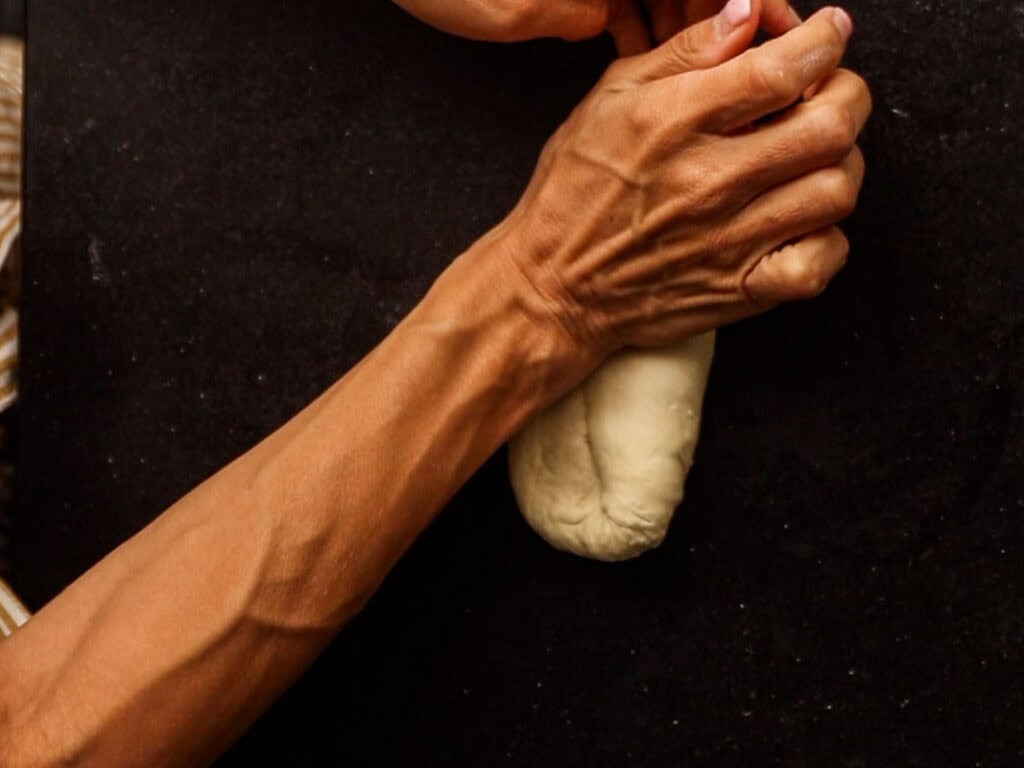 How to Make Delicious NY Style Bagels - Step by Step Guide. 28 A person kneads a small loaf of dough—reminiscent of traditional NY style bagels—on a dark surface, with their hands and forearms visible.
