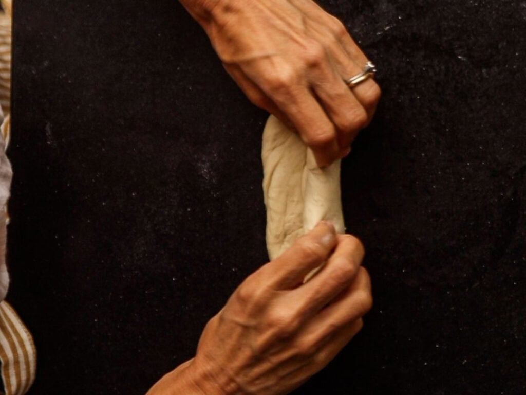 How to Make Delicious NY Style Bagels - Step by Step Guide. 29 Two hands knead and shape a piece of dough on a dark surface. The person, wearing a ring on one hand, expertly stretches the dough into a narrow shape—perfect for forming authentic NY style bagels.