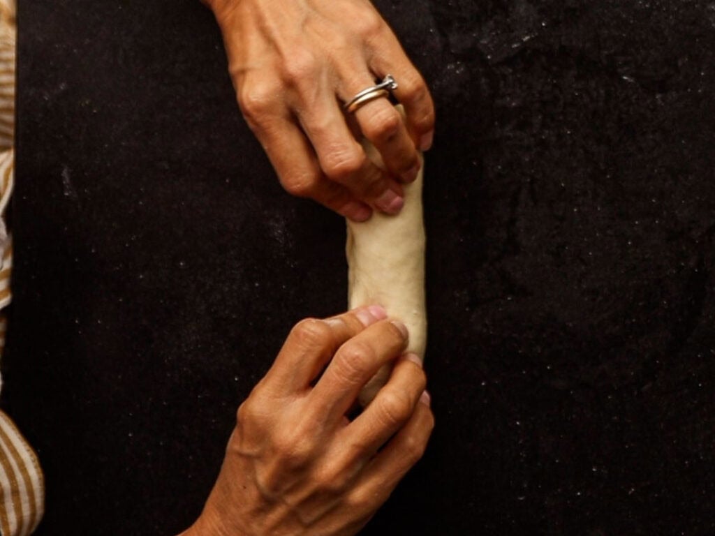 How to Make Delicious NY Style Bagels - Step by Step Guide. 30 Two hands roll a small piece of dough on a dark surface, shaping it into a log—just like preparing classic NY style bagels. The person wears a striped sleeve and a silver ring on their finger.