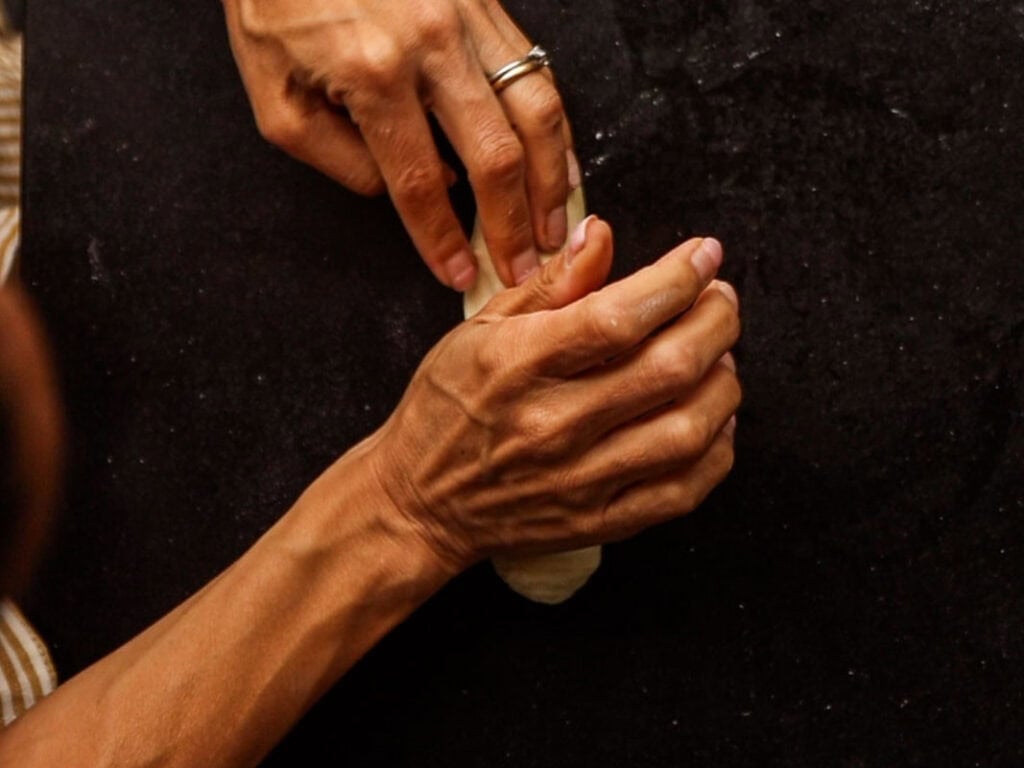 How to Make Delicious NY Style Bagels - Step by Step Guide. 31 Two hands, one adorned with a silver ring, are shaping or rolling a piece of dough on a dark surface—capturing the classic technique used for making authentic NY style bagels.
