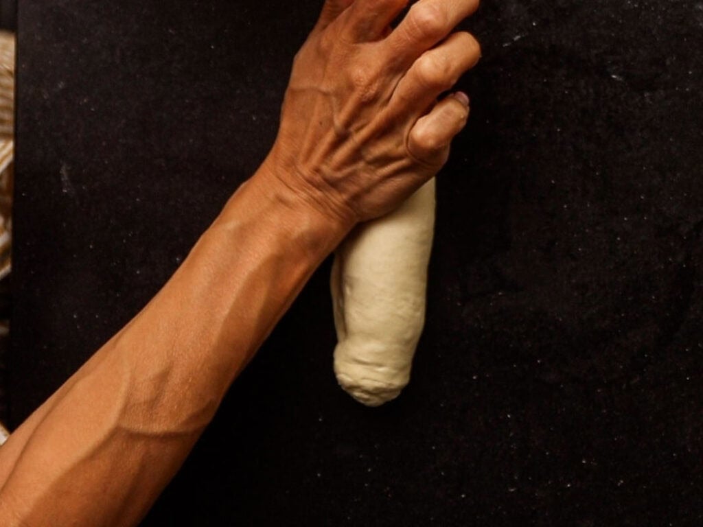 How to Make Delicious NY Style Bagels - Step by Step Guide. 32 A person’s hand shapes a piece of dough on a dark surface, carefully preparing it for baking authentic NY style bagels.