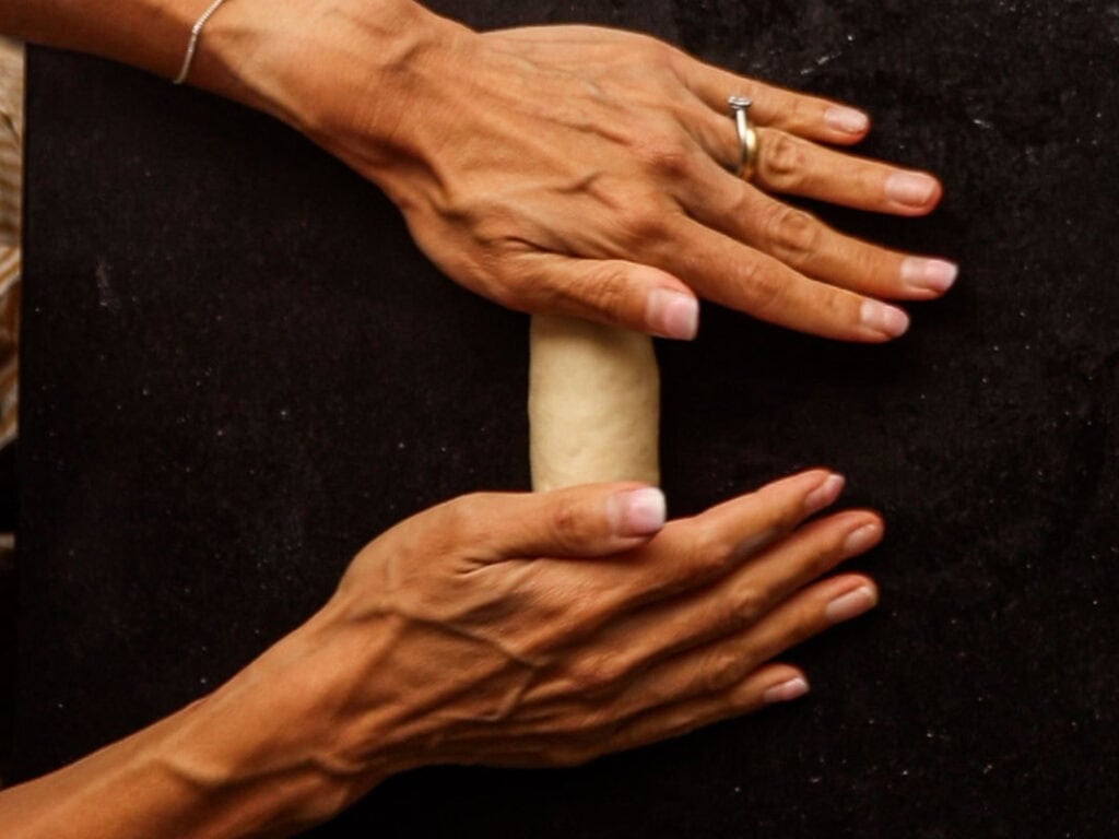 How to Make Delicious NY Style Bagels - Step by Step Guide. 34 Two hands with a ring on one finger are shaping or rolling a piece of dough on a dark surface, carefully forming it into the classic round shape of NY style bagels.