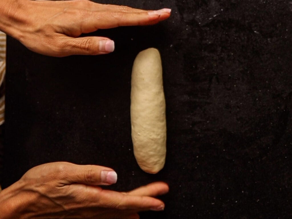 How to Make Delicious NY Style Bagels - Step by Step Guide. 35 Two hands shape a small, oval piece of dough on a dark surface, skillfully preparing it for baking into authentic NY style bagels.