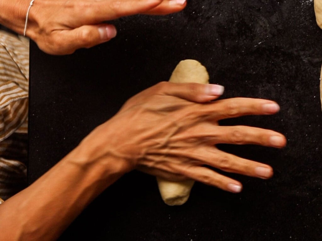 How to Make Delicious NY Style Bagels - Step by Step Guide. 36 Two hands are rolling dough on a dark surface, expertly shaping what will become classic NY style bagels. One hand is at the top while the other forms the dough in the center, with glimpses of striped clothing and the floured surface partially visible.
