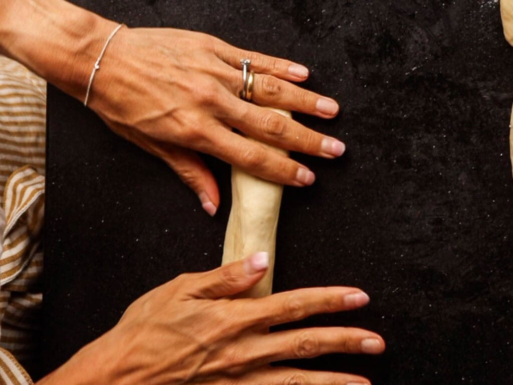 How to Make Delicious NY Style Bagels - Step by Step Guide. 37 Two hands roll a piece of dough on a dark surface, shaping it into a log—just the first step in making authentic NY style bagels. A striped cloth is partially visible on the left side.