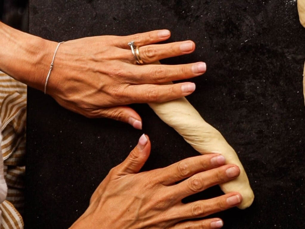 How to Make Delicious NY Style Bagels - Step by Step Guide. 38 Two hands are rolling a piece of dough into a long shape on a dark countertop, preparing it for classic NY style bagels. One hand wears a silver ring and bracelet, while part of a striped sleeve is visible on the left.