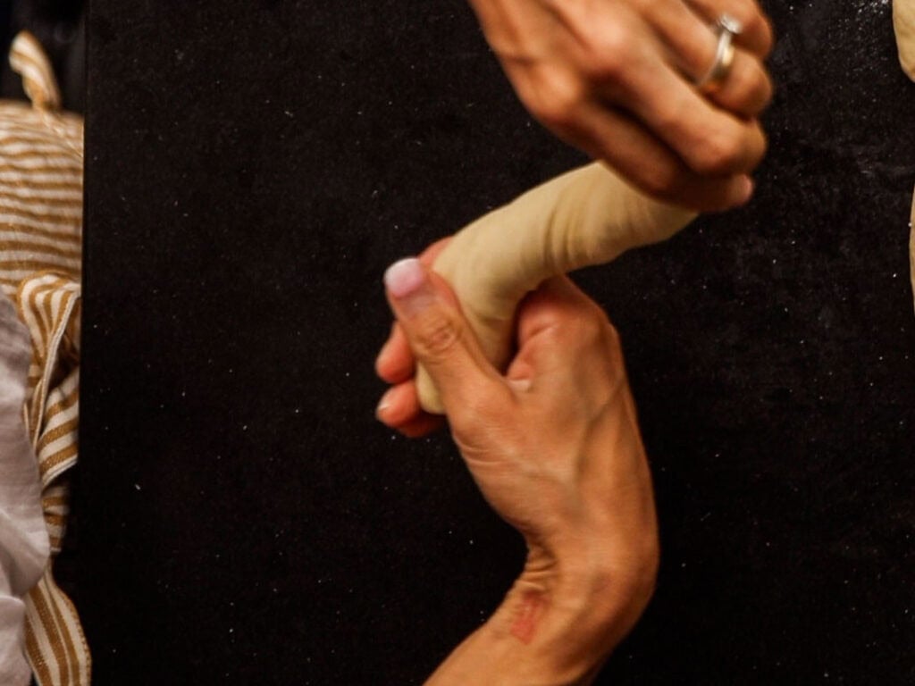 How to Make Delicious NY Style Bagels - Step by Step Guide. 39 Two hands roll a piece of dough on a dark surface, preparing it for classic ny style bagels. One hand wears a ring, and a person in striped clothing is partially visible on the left side.