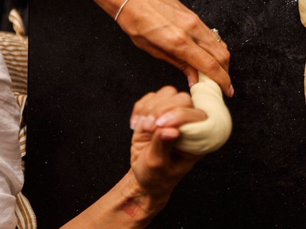 How to Make Delicious NY Style Bagels - Step by Step Guide. 40 Two hands twist a rope of dough against a dark surface, preparing to shape it into classic NY style bagels. One hand wears a thin silver bracelet. The process appears to be part of bread or pastry making.