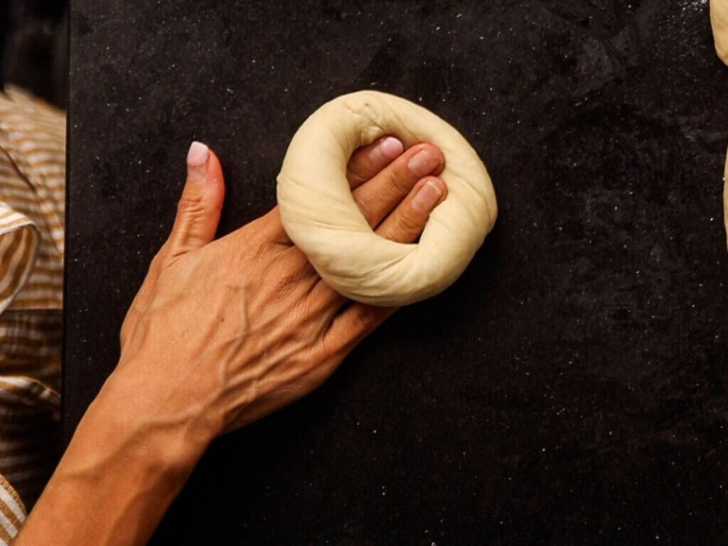 How to Make Delicious NY Style Bagels - Step by Step Guide. 48 A hand shaping dough into a ring on a dark surface, preparing to make classic NY style bagels.