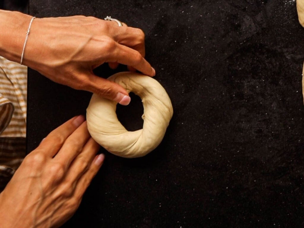 How to Make Delicious NY Style Bagels - Step by Step Guide. 49 A person shapes a ring of dough on a dark surface, preparing authentic NY style bagels for baking.