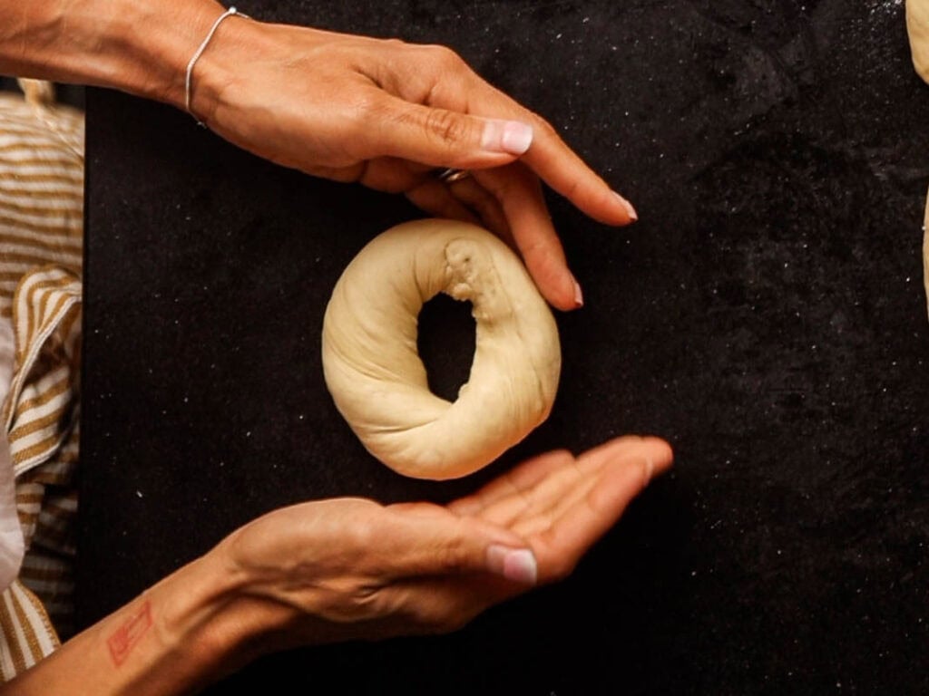 How to Make Delicious NY Style Bagels - Step by Step Guide. 50 Two hands shape a round piece of dough with a hole in the center on a dark surface, expertly forming what will become classic NY style bagels. A striped cloth is partially visible at the edge of the image.