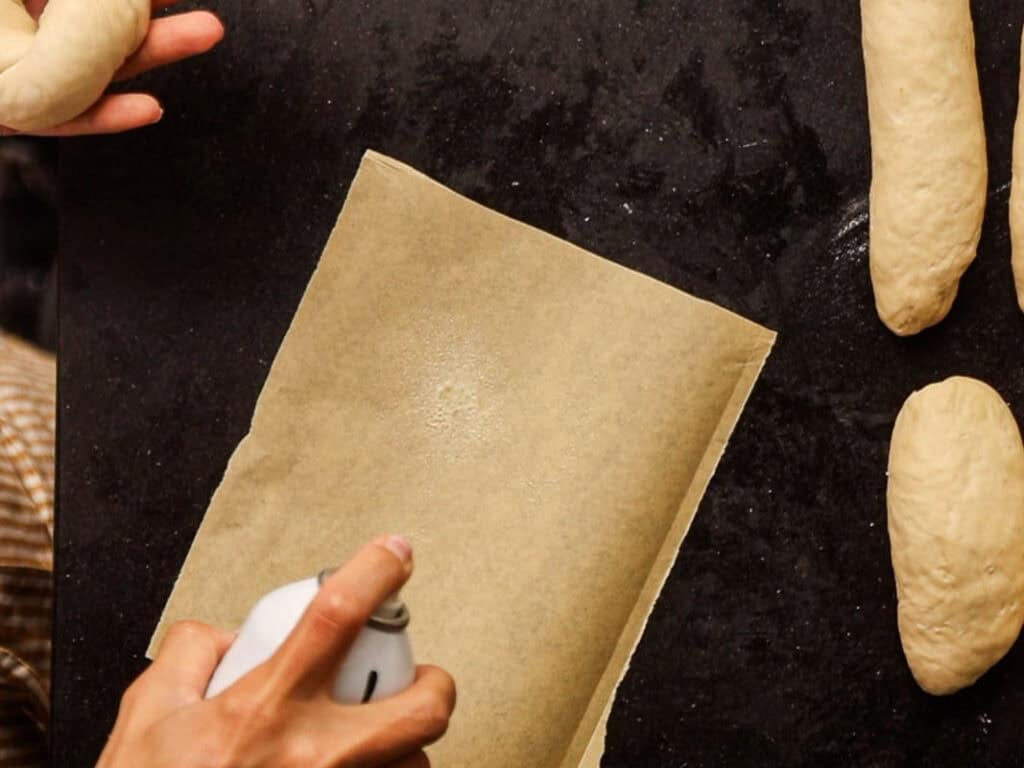 How to Make Delicious NY Style Bagels - Step by Step Guide. 51 A person sprays parchment paper with oil beside several pieces of raw NY style bagels dough on a dark countertop.
