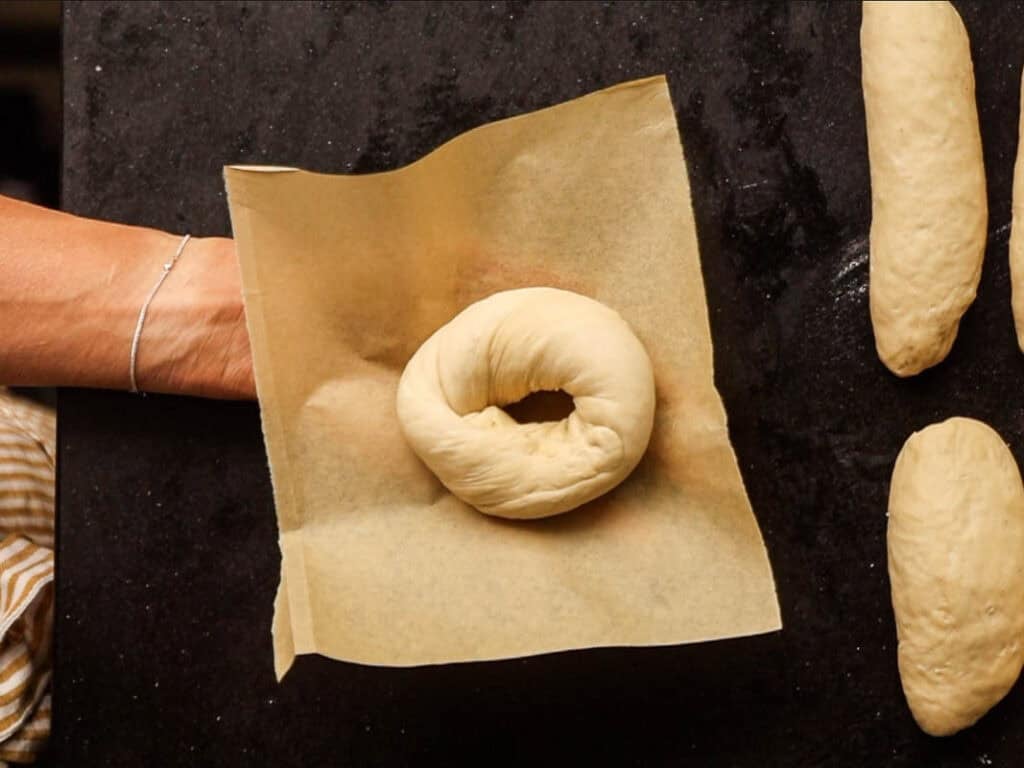 How to Make Delicious NY Style Bagels - Step by Step Guide. 52 A hand holds a piece of parchment paper with an unbaked NY style bagel dough on it, above a dark surface with more dough pieces nearby.