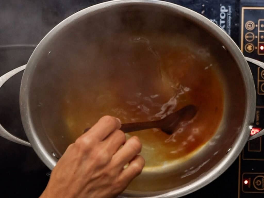 How to Make Delicious NY Style Bagels - Step by Step Guide. 57 A hand stirs a steaming pot of brown liquid with a wooden spoon on a stovetop, preparing the perfect base for authentic NY style bagels, with control buttons visible on the right side.
