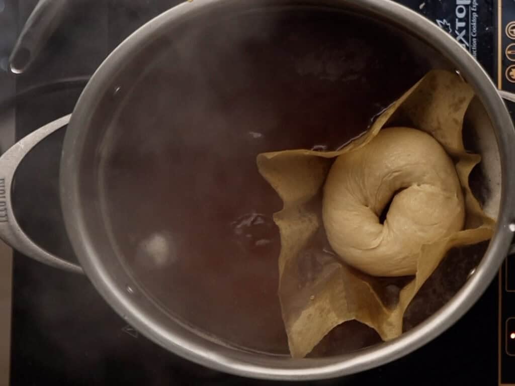 How to Make Delicious NY Style Bagels - Step by Step Guide. 60 A classic NY style bagel on parchment paper is being boiled in a pot of water on a stovetop, with steam rising from the pot.