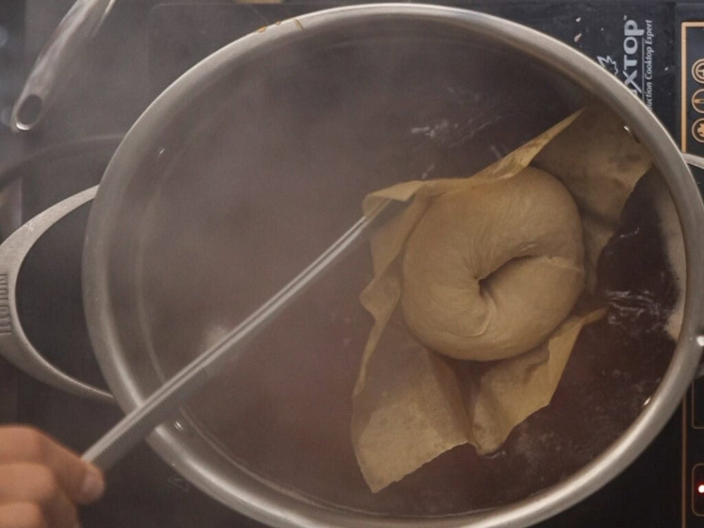 How to Make Delicious NY Style Bagels - Step by Step Guide. 61 A hand lowers a raw NY style bagel on parchment paper into a pot of boiling water on a stovetop, with steam rising around.