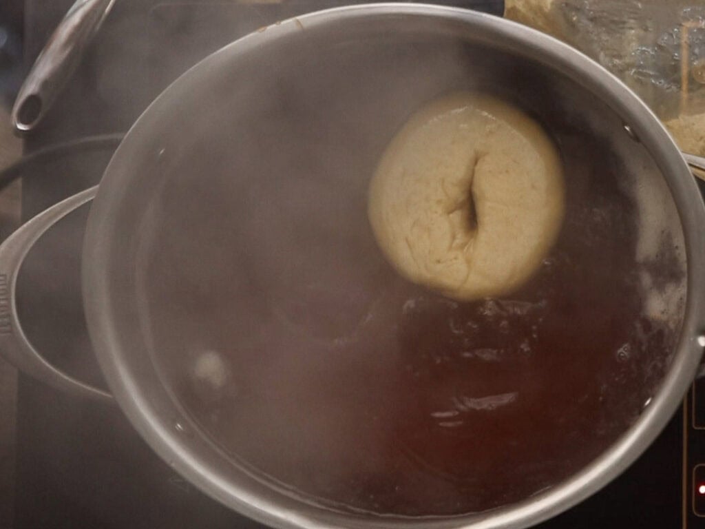How to Make Delicious NY Style Bagels - Step by Step Guide. 62 A single NY style bagel is boiling in a large pot of water on a stovetop, with steam rising from the pot.