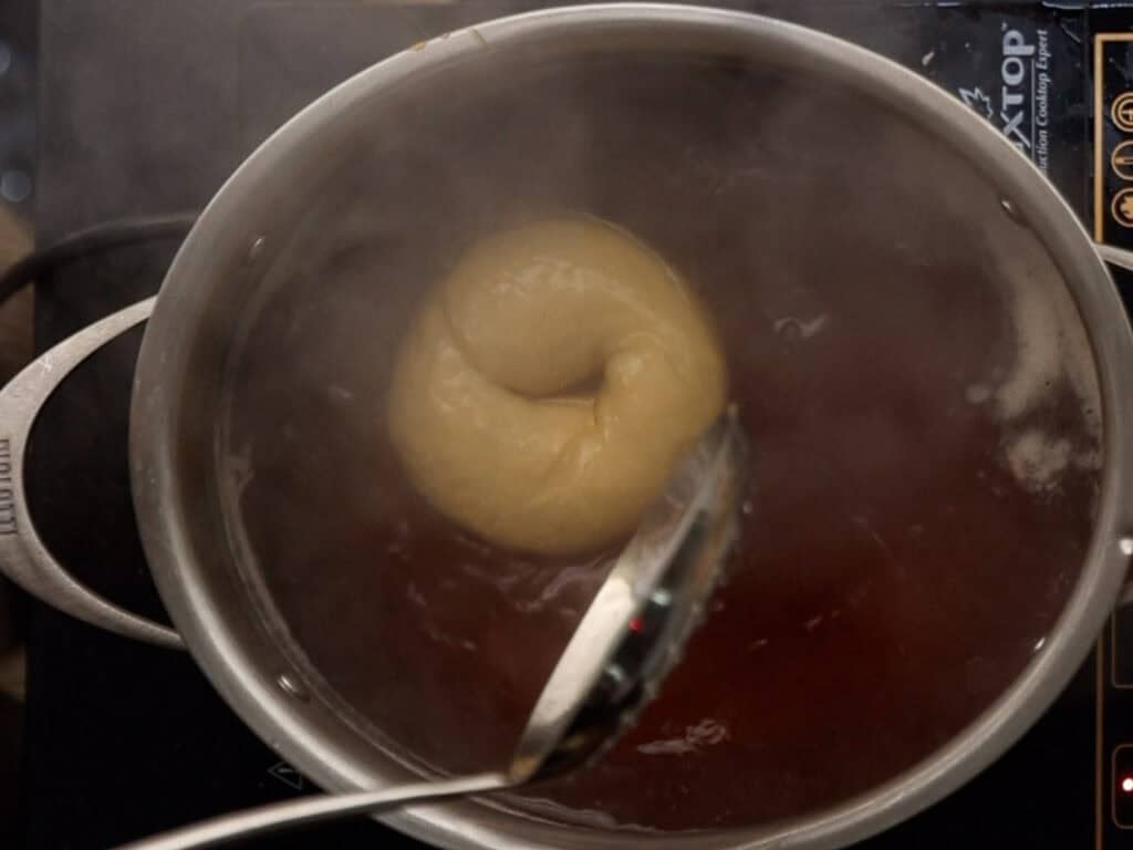 How to Make Delicious NY Style Bagels - Step by Step Guide. 63 A classic NY style bagel is being boiled in a pot of water on a stovetop, with steam rising and a slotted spoon nearby.