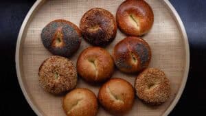 A round tray holds nine NY style bagels, some topped with seeds like poppy and sesame, and others plain. The bagels have a golden-brown crust and are arranged in a loose grid.