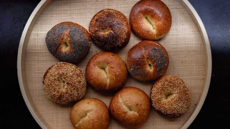 A round tray holds nine NY style bagels, some topped with seeds like poppy and sesame, and others plain. The bagels have a golden-brown crust and are arranged in a loose grid.