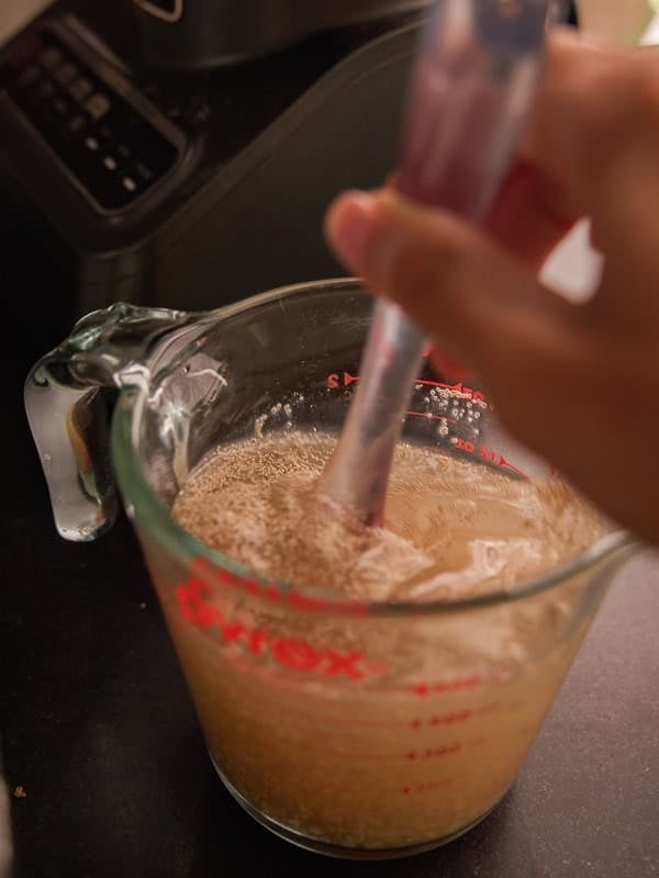 How to Make Delicious NY Style Bagels - Step by Step Guide. 17 A hand stirs a cloudy liquid with visible yeast in a glass measuring cup on a kitchen counter, preparing the base for authentic NY style bagels.