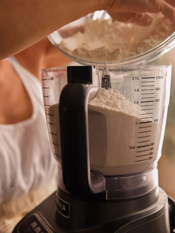 How to Make Delicious NY Style Bagels - Step by Step Guide. 18 A person pours flour from a bowl into a food processor filled with flour, preparing dough for classic NY style bagels. The food processor has measurement markings on the side.