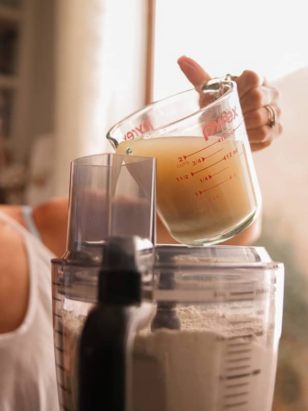 How to Make Delicious NY Style Bagels - Step by Step Guide. 19 A person pours liquid from a measuring cup into a food processor containing flour, preparing dough for authentic NY style bagels.