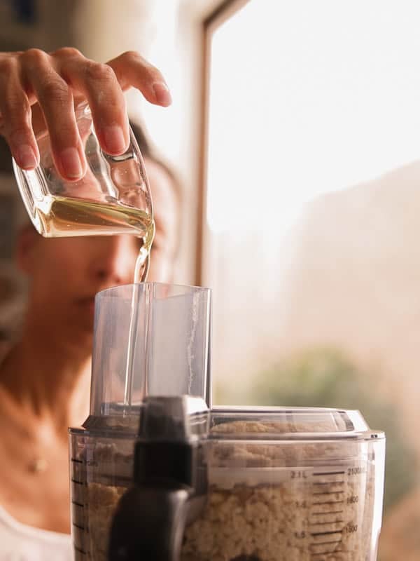 How to Make Delicious NY Style Bagels - Step by Step Guide. 20 A person pours oil from a small glass container into a food processor filled with chopped ingredients for homemade NY style bagels, with soft natural light in the background.