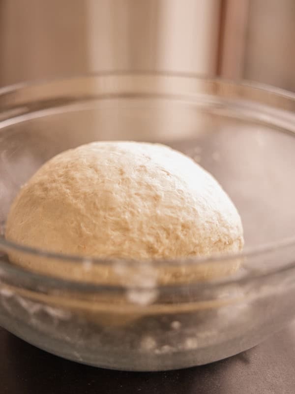 How to Make Delicious NY Style Bagels - Step by Step Guide. 23 A ball of dough for NY style bagels rises in a clear glass bowl, placed on a dark surface, with soft light coming from the background.