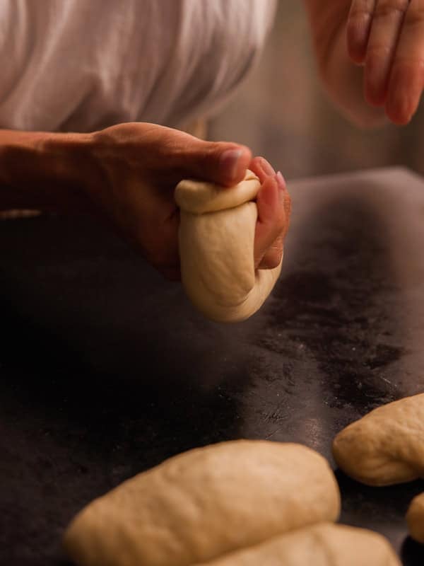 How to Make Delicious NY Style Bagels - Step by Step Guide. 41 A person shapes a piece of dough by hand on a dark surface, with other dough pieces nearby, suggesting the preparation of NY style bagels or similar bread treats.