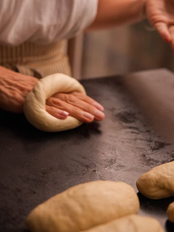 How to Make Delicious NY Style Bagels - Step by Step Guide. 44 A person shapes dough into a ring on a dark surface, preparing classic ny style bagels. Several pieces of unshaped dough rest nearby. Only the hands and part of the torso are visible.