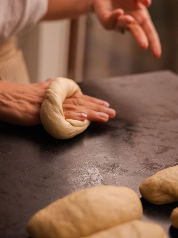 How to Make Delicious NY Style Bagels - Step by Step Guide. 45 Hands shaping dough on a dark countertop, preparing it for baking. Several pieces of dough are visible in the foreground, suggesting a classic ny style bagels baking or bread-making process.