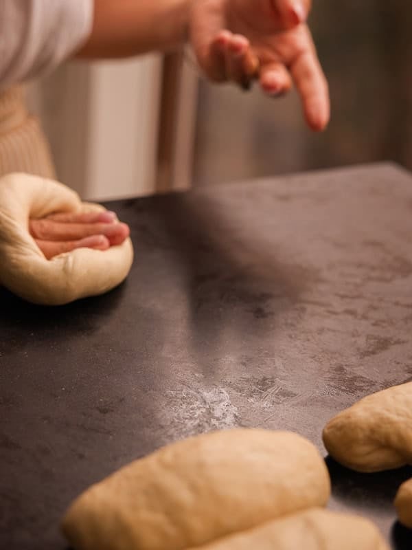How to Make Delicious NY Style Bagels - Step by Step Guide. 47 A person shapes dough by hand on a dark countertop, preparing several pieces for baking authentic NY style bagels; only their hands and some dough are visible.