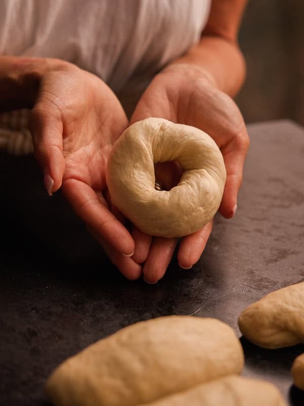 How My Bagels Triggered The Sexist In Me. 4 A person holds an unbaked, round ny style bagel dough in their hands over a dark surface with other dough pieces nearby.