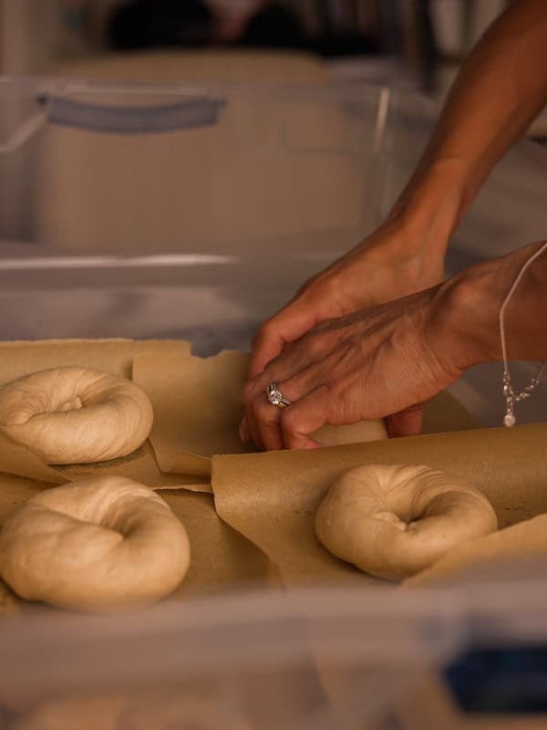 How to Make Delicious NY Style Bagels - Step by Step Guide. 53 A person’s hands shape raw ny style bagel dough on parchment paper in a tray, preparing the bagels for baking.