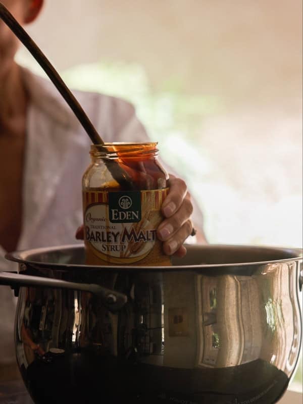 How to Make Delicious NY Style Bagels - Step by Step Guide. 15 A person holds a jar of Eden Barley Malt Syrup above a stainless steel pot, using a spoon to scoop syrup—the classic secret ingredient for making authentic NY style bagels. The background is softly blurred.