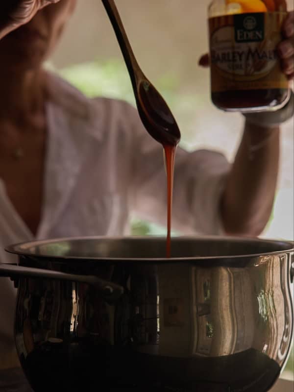 How My Bagels Triggered The Sexist In Me. 3 A person pours barley malt syrup from a wooden spoon into a metal pot, holding a jar labeled Eden Barley Malt—the secret ingredient often used for authentic NY style bagels. The background is softly blurred.