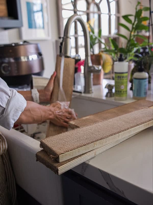How to Make Delicious NY Style Bagels - Step by Step Guide. 11 A person washes long, rectangular fabric-covered boards in a kitchen sink—preparing them perhaps for the next batch of freshly baked NY style bagels. Two clean boards rest on the counter nearby, while plants, soap, and other kitchen items are visible in the background.