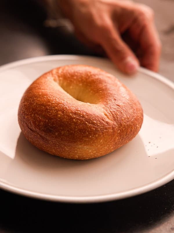 How to Make Delicious NY Style Bagels - Step by Step Guide. 3 A plain, golden-brown NY style bagel sits on a white plate, with a hand reaching toward it from the top right corner of the image.