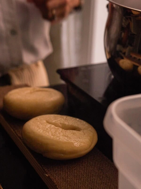How to Make Delicious NY Style Bagels - Step by Step Guide. 65 Two unbaked, round NY style bagels rest on a brown tray near a metal pot and a plastic container, with a person standing in the background.