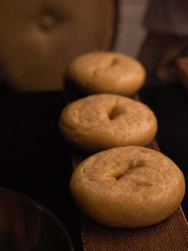 How to Make Delicious NY Style Bagels - Step by Step Guide. 69 Three plain NY style bagels are arranged in a row on a brown textured surface, with a cushioned chair and a dark background visible behind them.