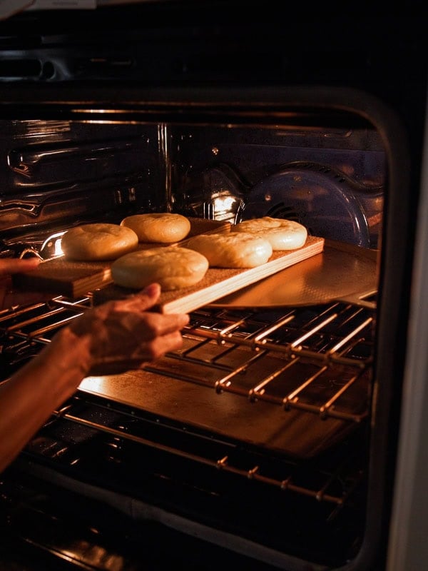 How My Bagels Triggered The Sexist In Me. 5 A person places a wooden tray with six round dough pieces—resembling classic NY style bagels—into an oven to bake, with oven racks visible inside.