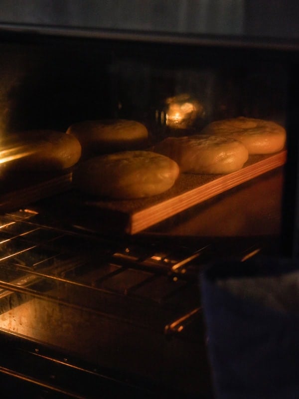 How to Make Delicious NY Style Bagels - Step by Step Guide. 71 Six round loaves of bread, reminiscent of classic NY style bagels, are baking on a wooden tray inside an oven, illuminated by the oven’s warm interior light.