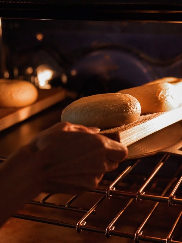 How to Make Delicious NY Style Bagels - Step by Step Guide. 72 A person uses a wooden peel to place dough onto an oven rack for baking, with other NY style bagels visible on a tray inside the oven.