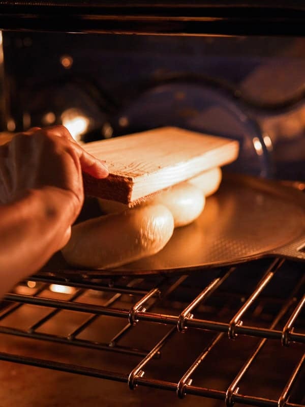 How to Make Delicious NY Style Bagels - Step by Step Guide. 74 A hand places a wooden board on top of dough rising on a baking tray inside an oven, preparing classic NY style bagels or bread for baking.