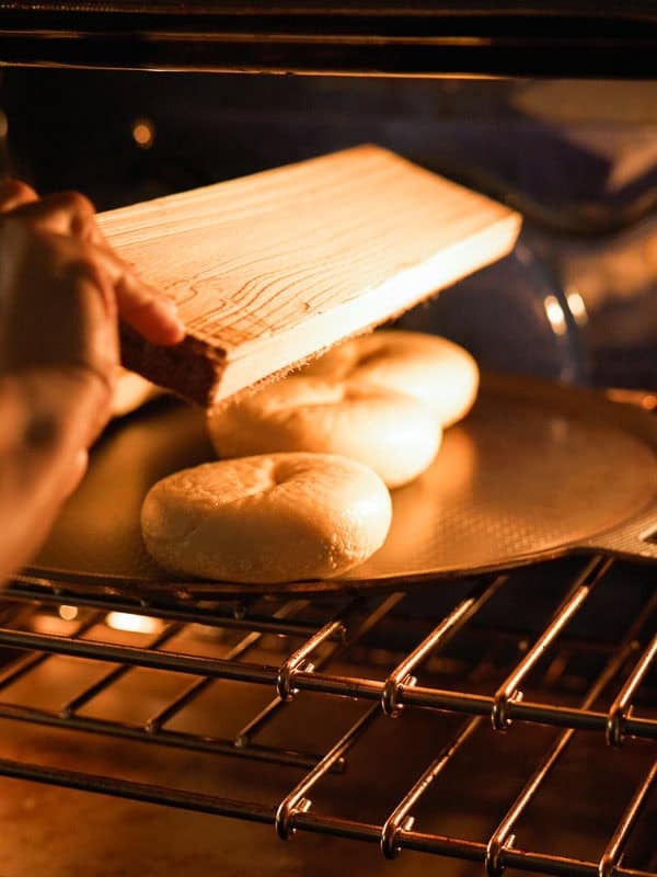 How to Make Delicious NY Style Bagels - Step by Step Guide. 75 A person slides raw NY style bagels from a wooden board onto a baking tray inside an oven. Warm light illuminates the bagels and the oven’s metal racks.