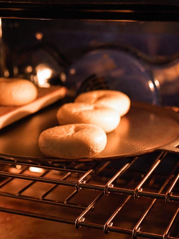 How to Make Delicious NY Style Bagels - Step by Step Guide. 76 Three round dough pieces, reminiscent of classic NY style bagels, are baking on a tray inside an oven, illuminated by the warm oven light.