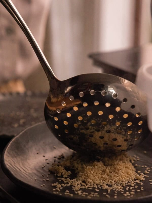 How to Make Delicious NY Style Bagels - Step by Step Guide. 66 A metal slotted spoon is scattering sesame seeds onto a dark plate, reminiscent of the classic topping found on NY style bagels. The background is softly blurred, highlighting the seeds and spoon.