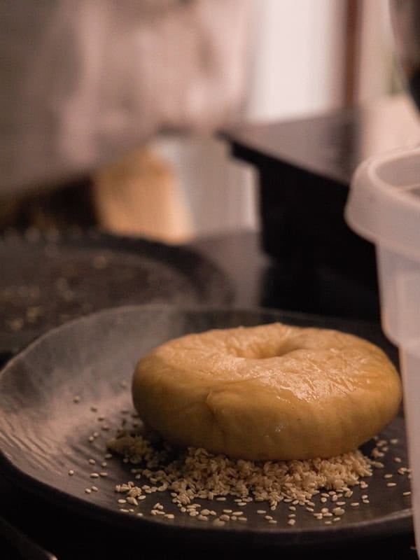 How to Make Delicious NY Style Bagels - Step by Step Guide. 67 A round, unbaked dough sits on a black plate, partially covered with sesame seeds—just waiting to become classic NY style bagels. A blurred kitchen background and a white container are visible.
