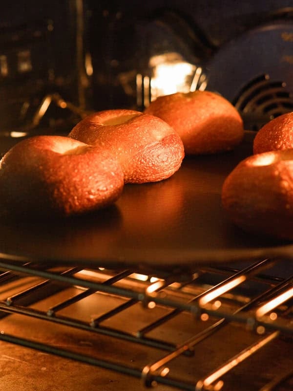 How to Make Delicious NY Style Bagels - Step by Step Guide. 77 Four NY style bagels baking on a tray inside an oven, with golden brown crusts visible under the warm oven light.